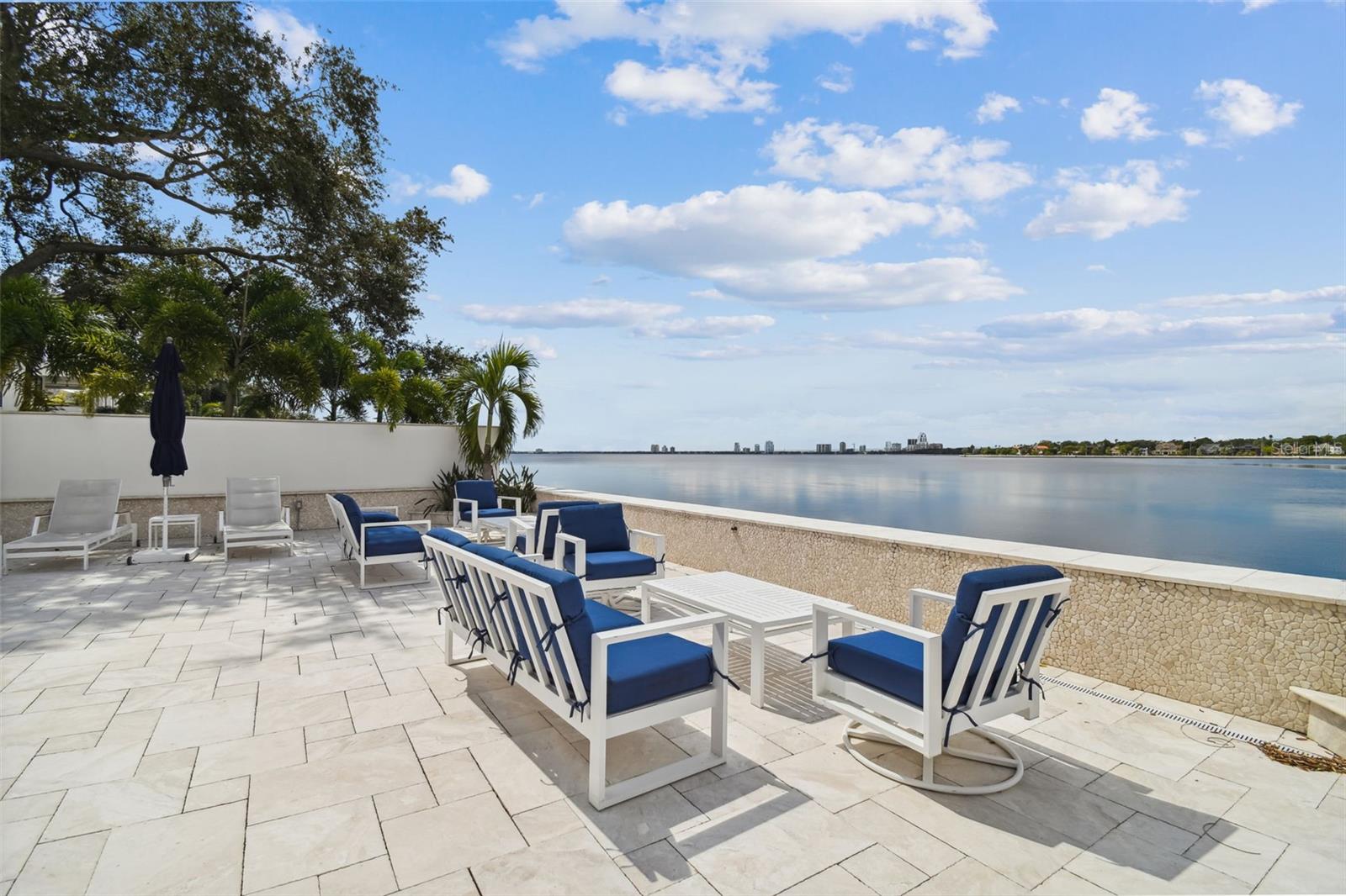 Waterfront terrace with blue-cushioned lounge chairs overlooking calm water and a distant skyline.