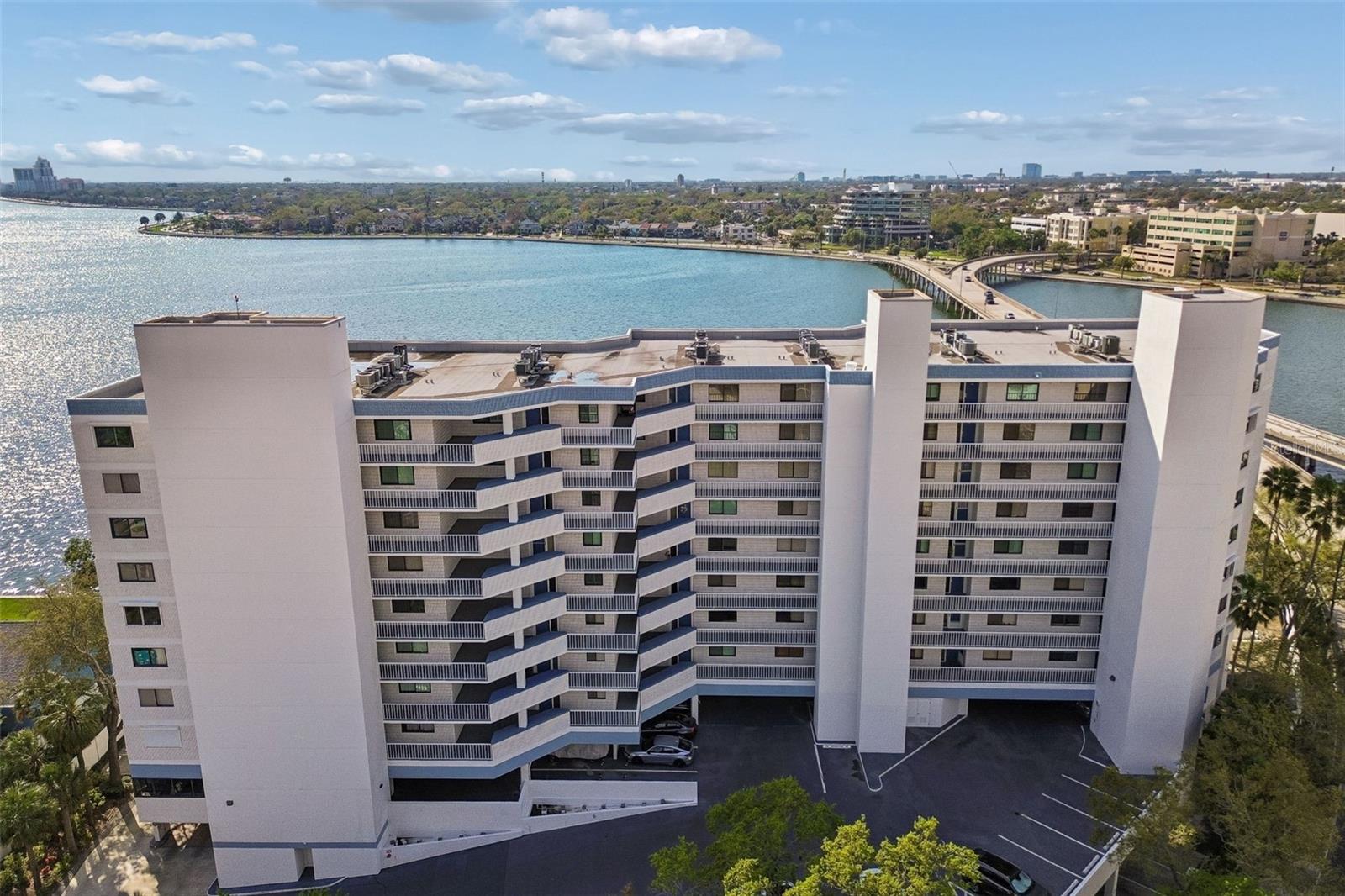 Aerial view of a waterfront condominium beside the bay, with a curving bridge and distant city buildings.