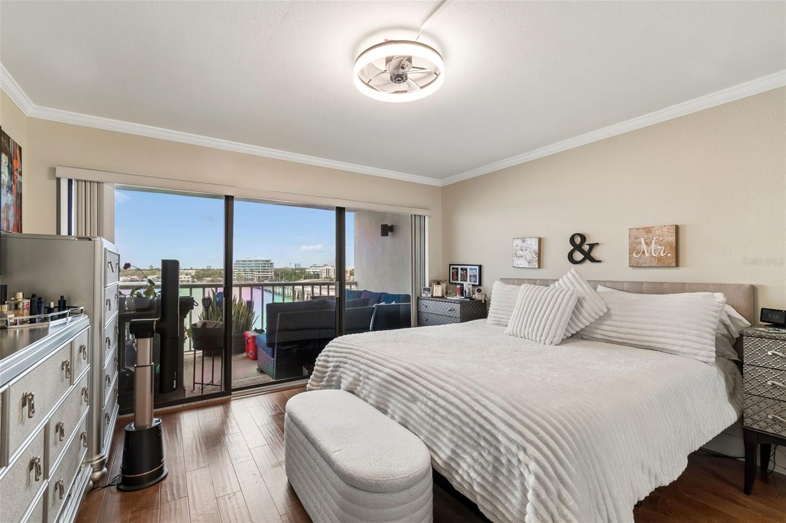 Bedroom with a white bed and balcony doors overlooking water and city buildings.