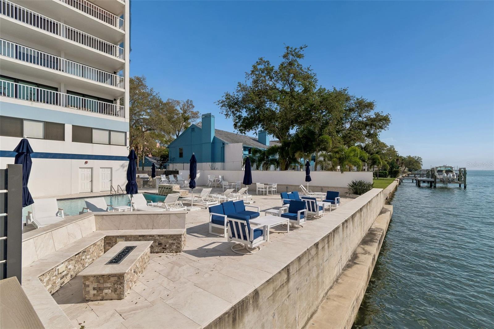 Condominium pool deck beside the water, with lounge chairs and a stone fire pit.