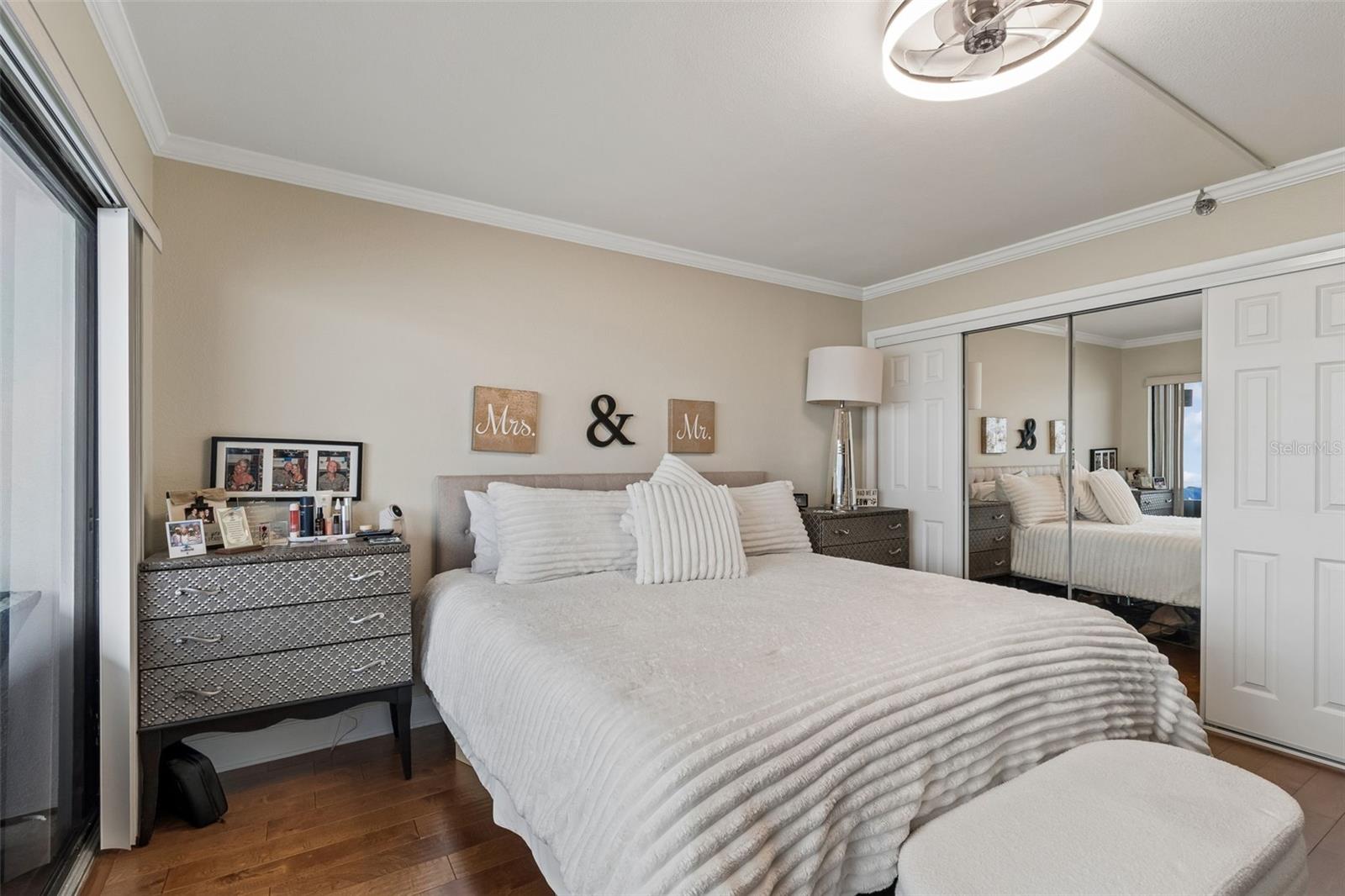 Furnished bedroom with a white bed, mirrored closet doors, and a patterned gray dresser.
