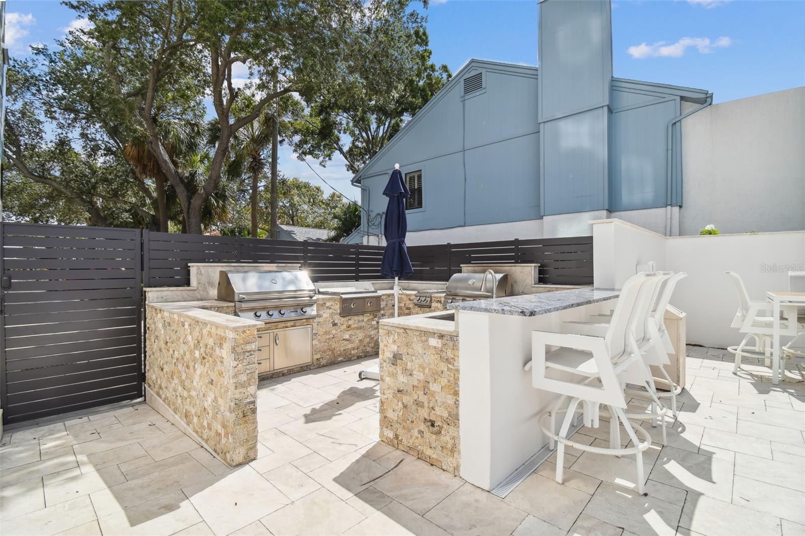 Outdoor kitchen patio with stainless steel grills, stone counters, white bar stools and a dark privacy fence.