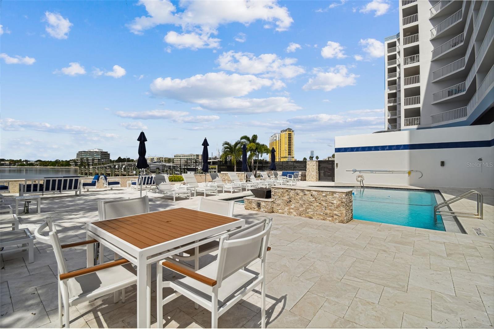 Condo pool deck with turquoise water, white loungers, patio seating, and a view across the water.