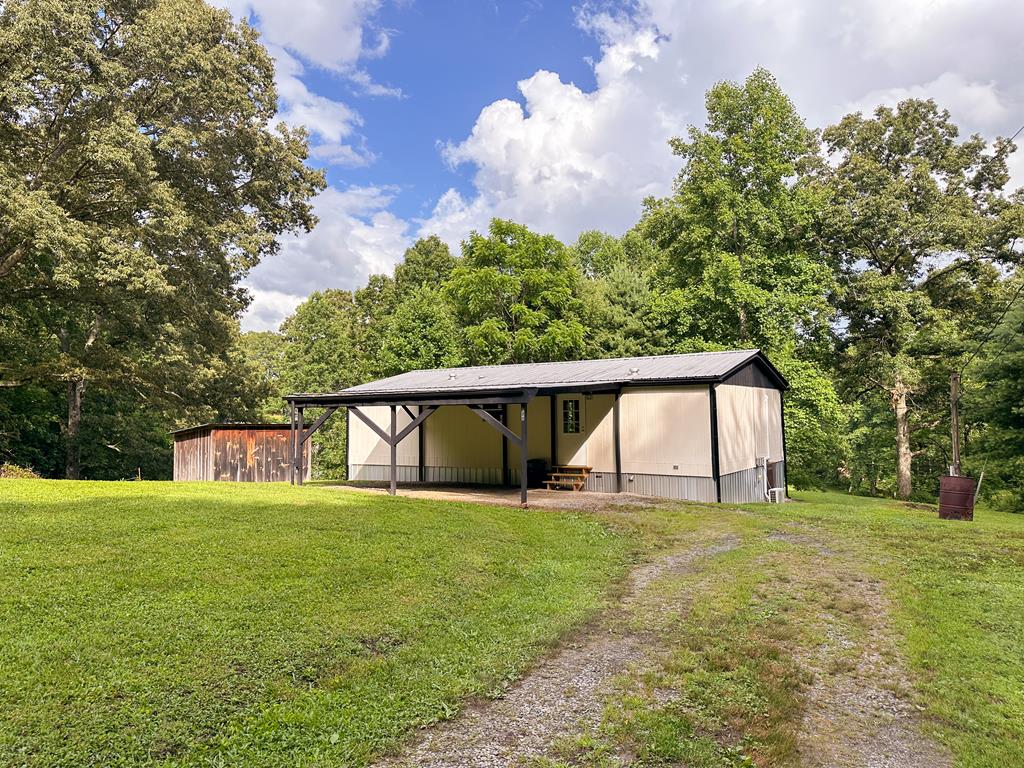 Metal outbuilding with an open-sided carport beside a weathered shed on a grassy, wooded lot.
