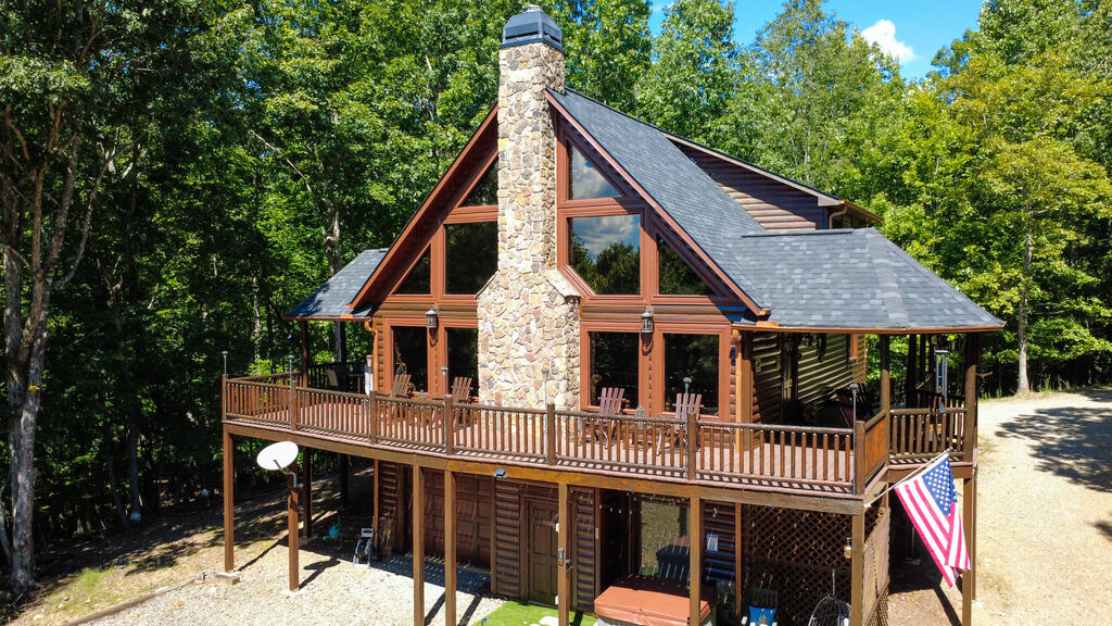 Rustic cabin with a stone chimney, large windows, and a raised wraparound deck in a wooded setting.