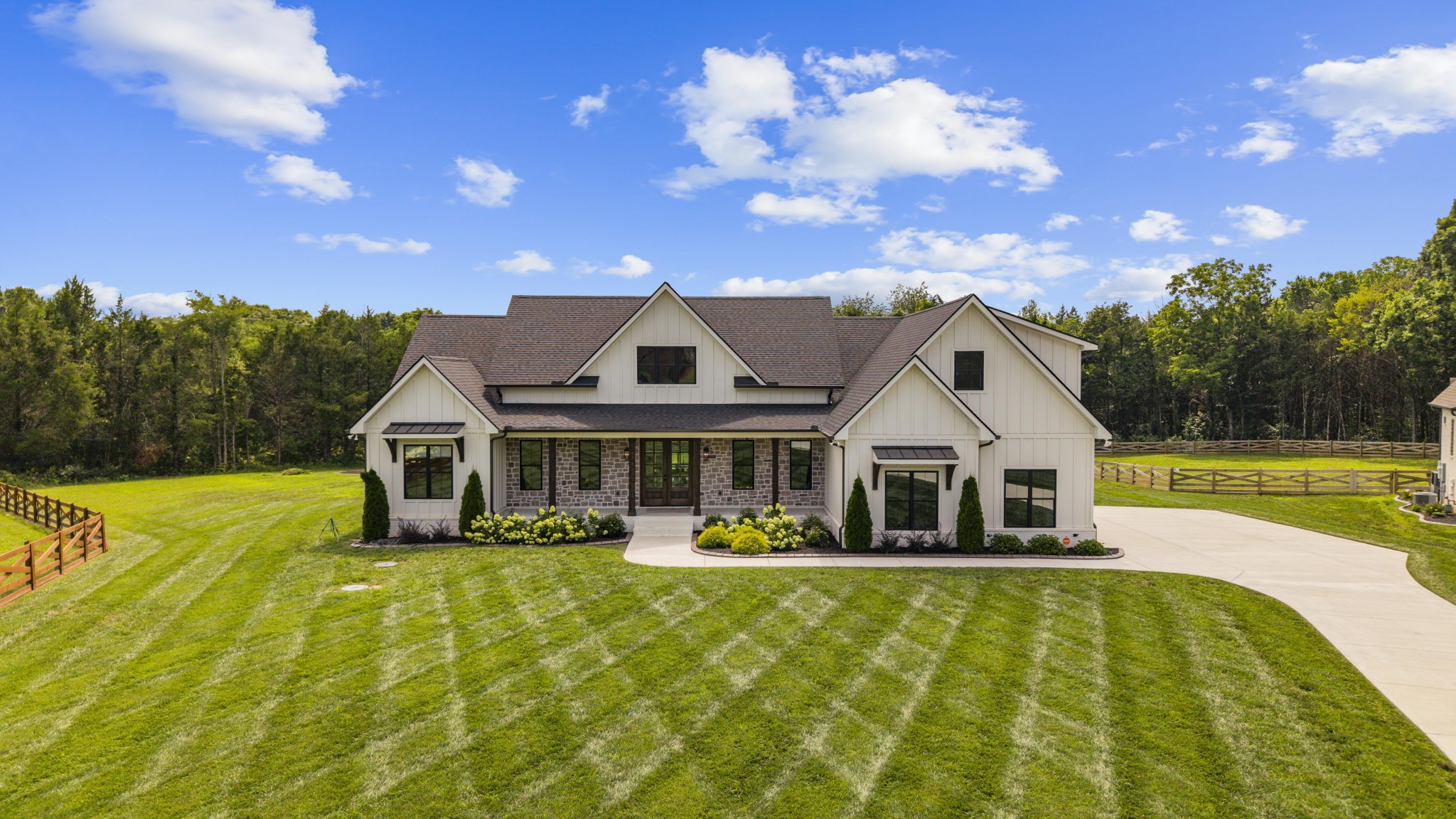 White-and-stone house with multiple gables, a broad lawn, wooden fencing, and woods behind it.