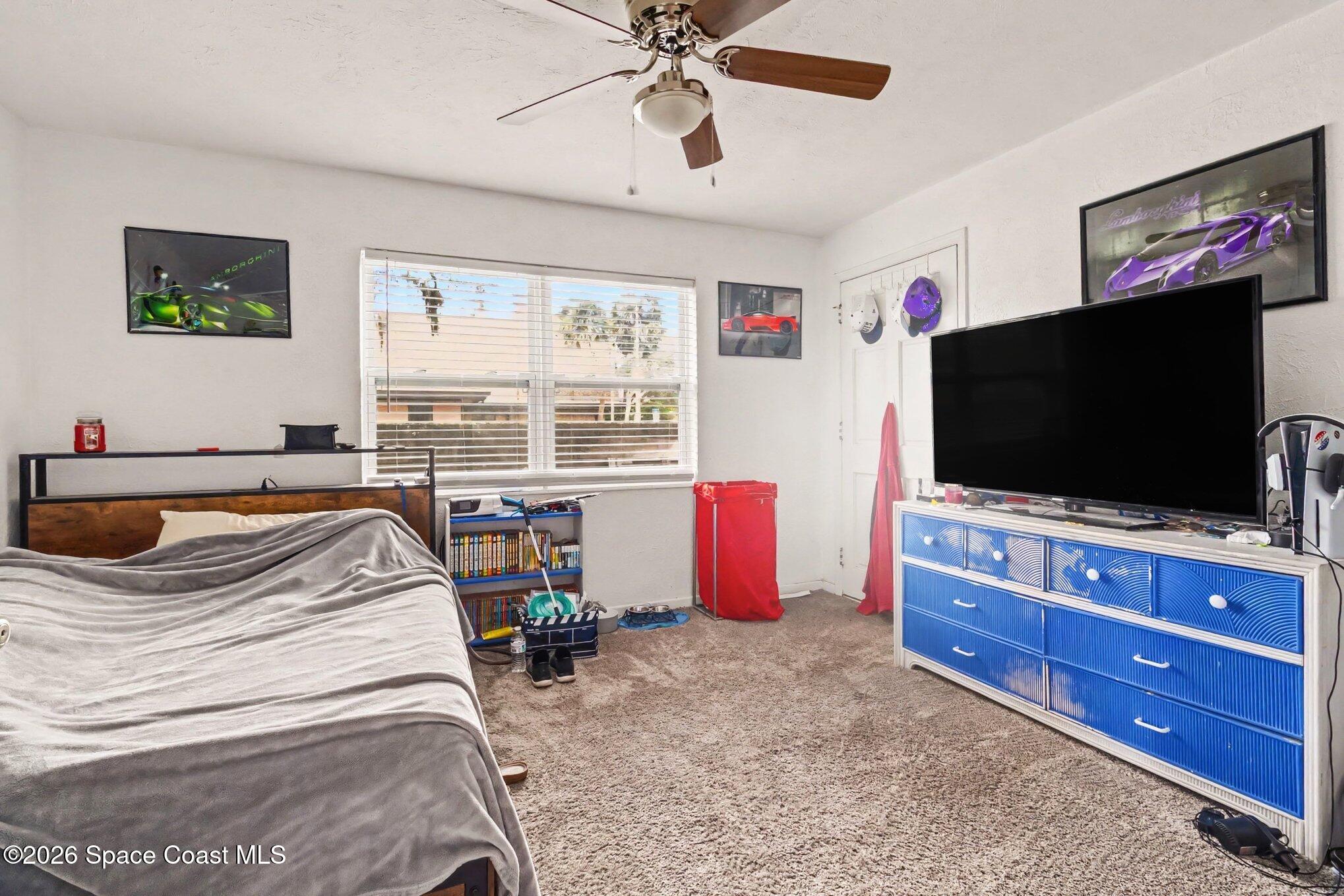 Bedroom with gray-covered bed, beige carpet, window blinds, and a television on a blue dresser.
