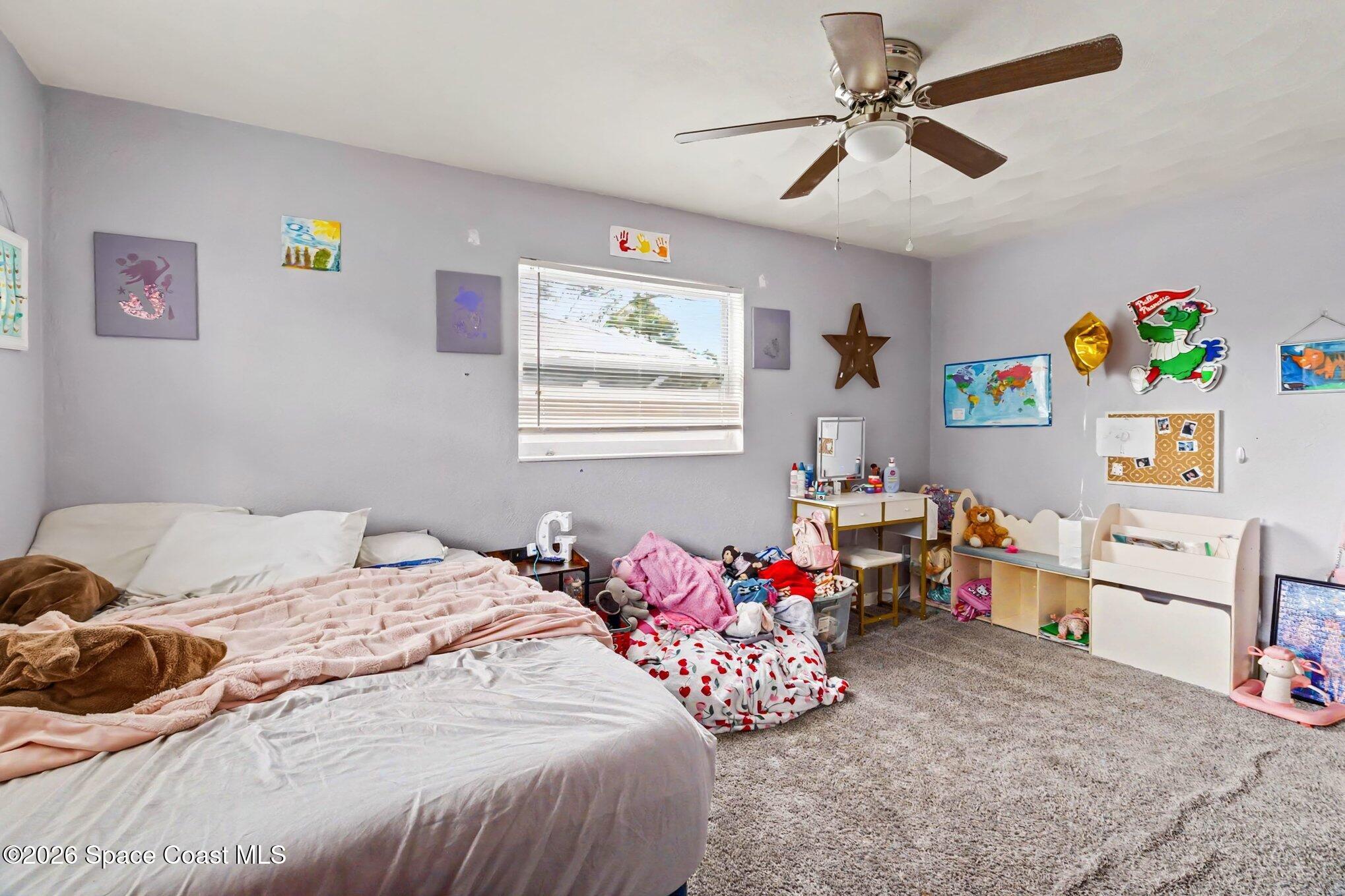 Bedroom with a large bed, gray carpet, children’s artwork, toys, and a ceiling fan.