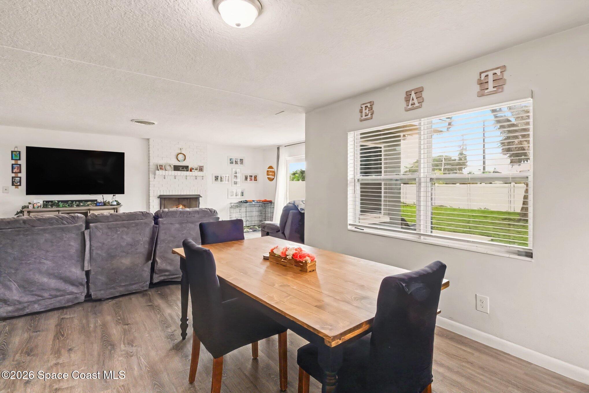 Dining area with a wooden table overlooks a living room with gray sofas and a white brick fireplace.