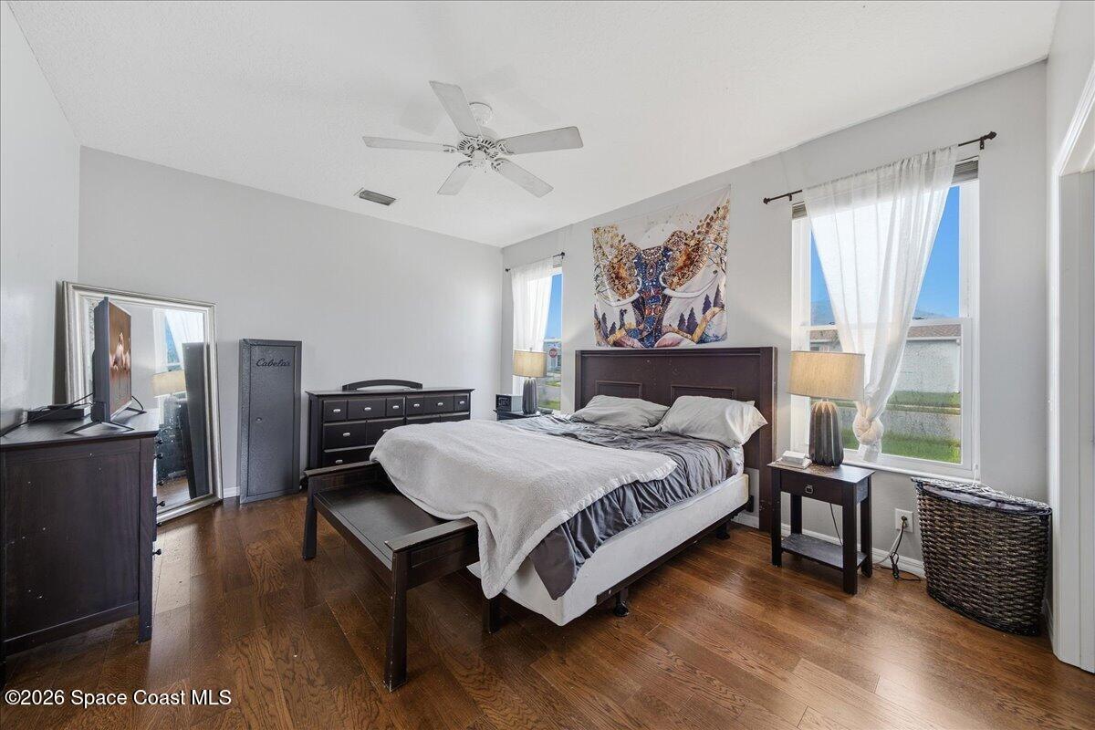 Furnished bedroom with a dark wood bed, hardwood floors, and two bright curtained windows.