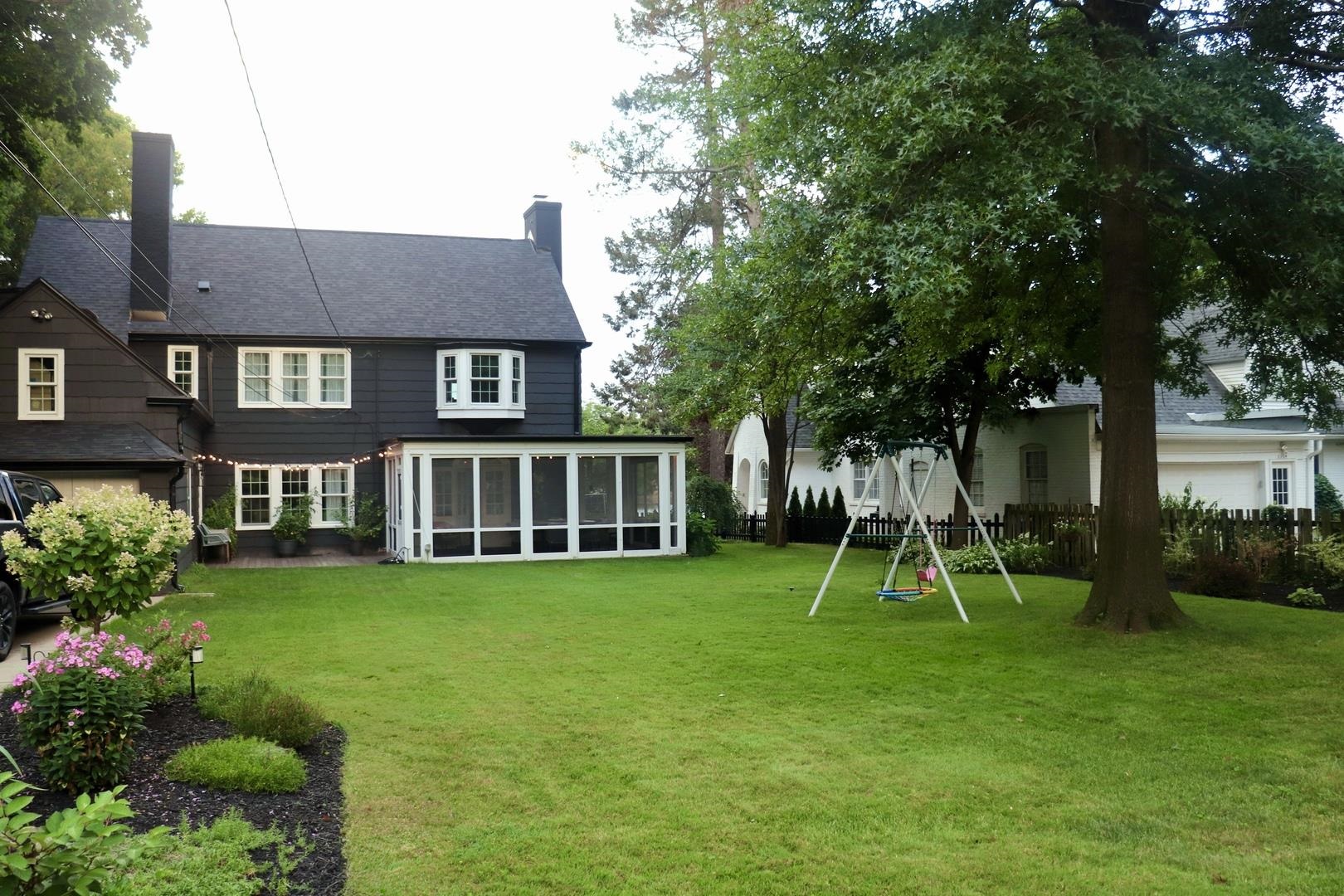 Rear of a dark two-story house with a screened porch, broad lawn, and swing set.