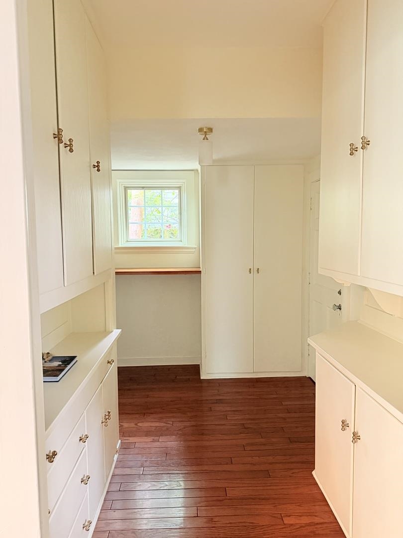 Walk-in closet with white built-in cabinetry, brass hardware, hardwood floors, and a window.