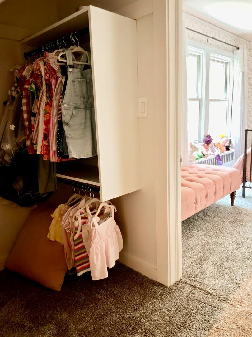 Open bedroom closet with hanging clothes beside a bright bedroom with a pink upholstered bed.