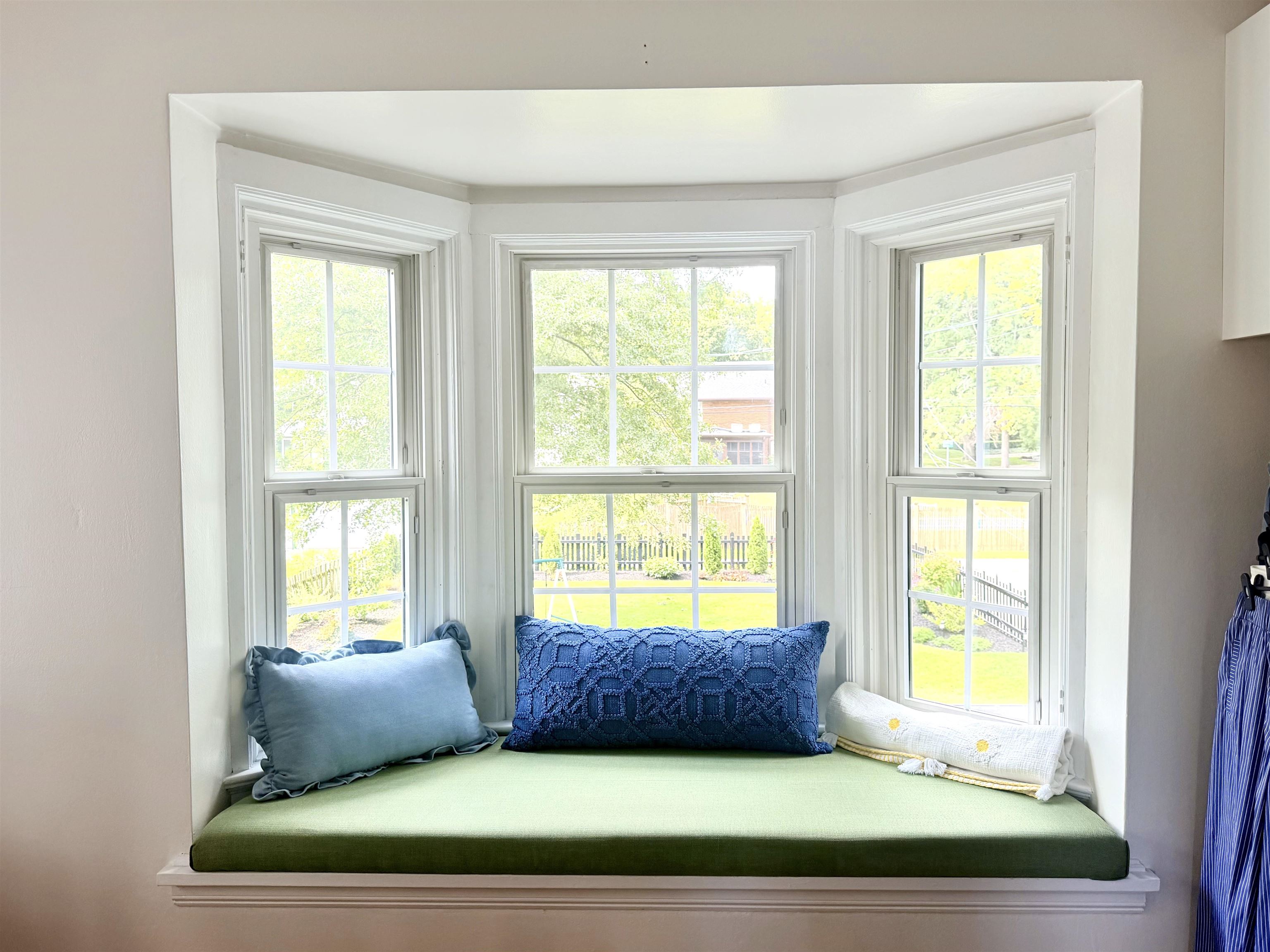 Built-in window seat with a green cushion and blue pillows beneath three bright windows overlooking a yard.