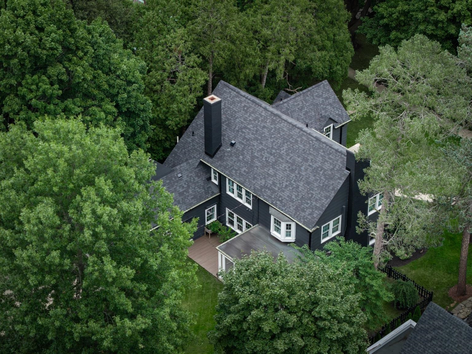 Aerial view of a dark-sided house with a gray shingle roof, deck, and mature trees.