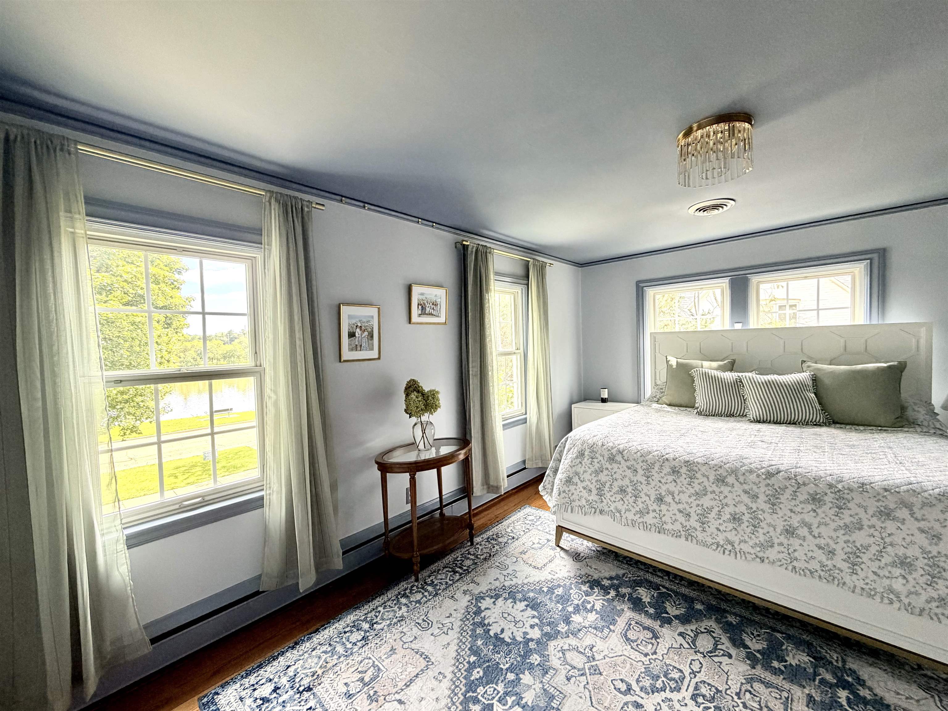 Bedroom with a large bed and patterned rug beside tall windows overlooking trees and water.