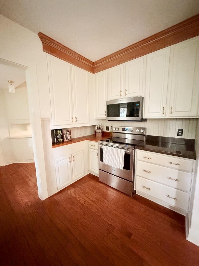 Kitchen with white paneled cabinets, stainless steel appliances, dark counters, and reddish hardwood flooring.