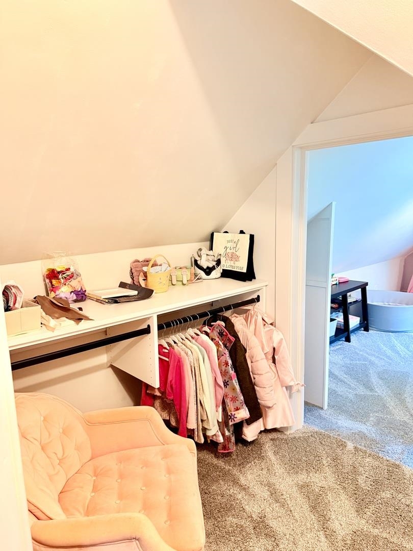 Dressing area with a clothing rack, storage shelf, and peach tufted loveseat beneath a sloped ceiling.