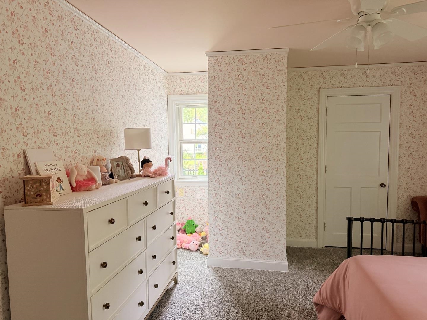 Bedroom with floral wallpaper, a white dresser topped with plush toys, and pink bedding.