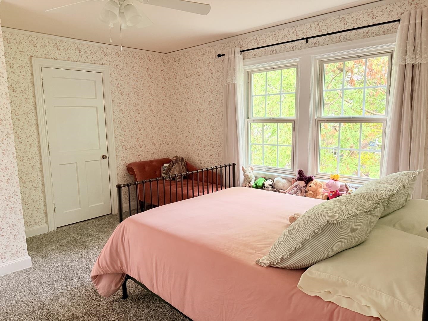 Bedroom with a pink-covered bed, floral wallpaper, and two bright windows beside a small loveseat.