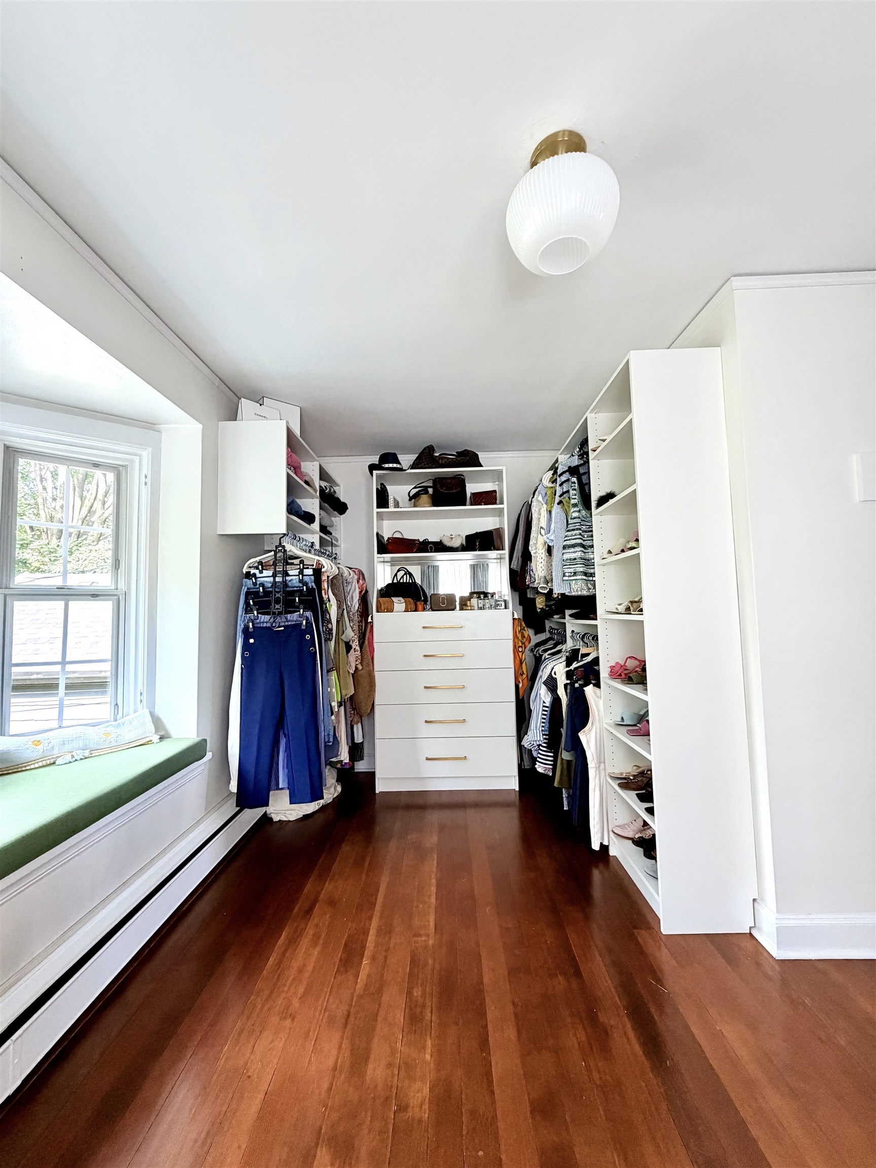 Walk-in closet with white built-in storage, hanging clothes, hardwood floors, and a window seat.