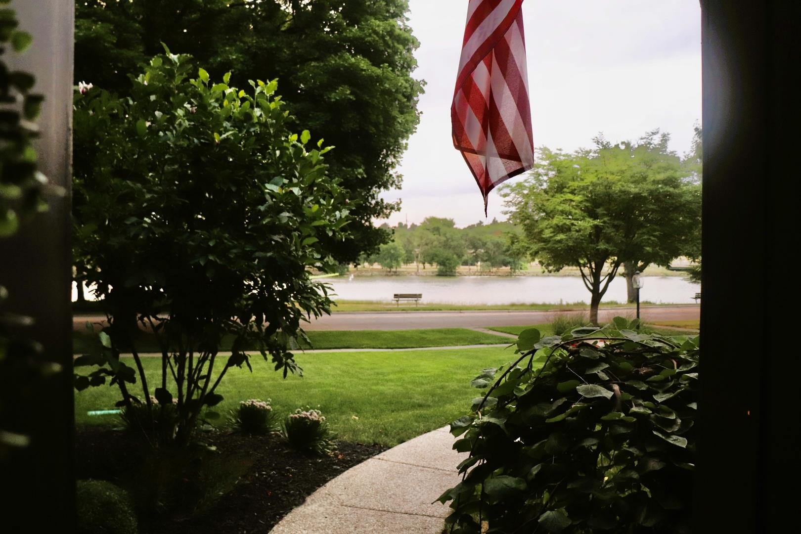 Front lawn and walkway overlook a lake, framed by mature trees and an American flag.