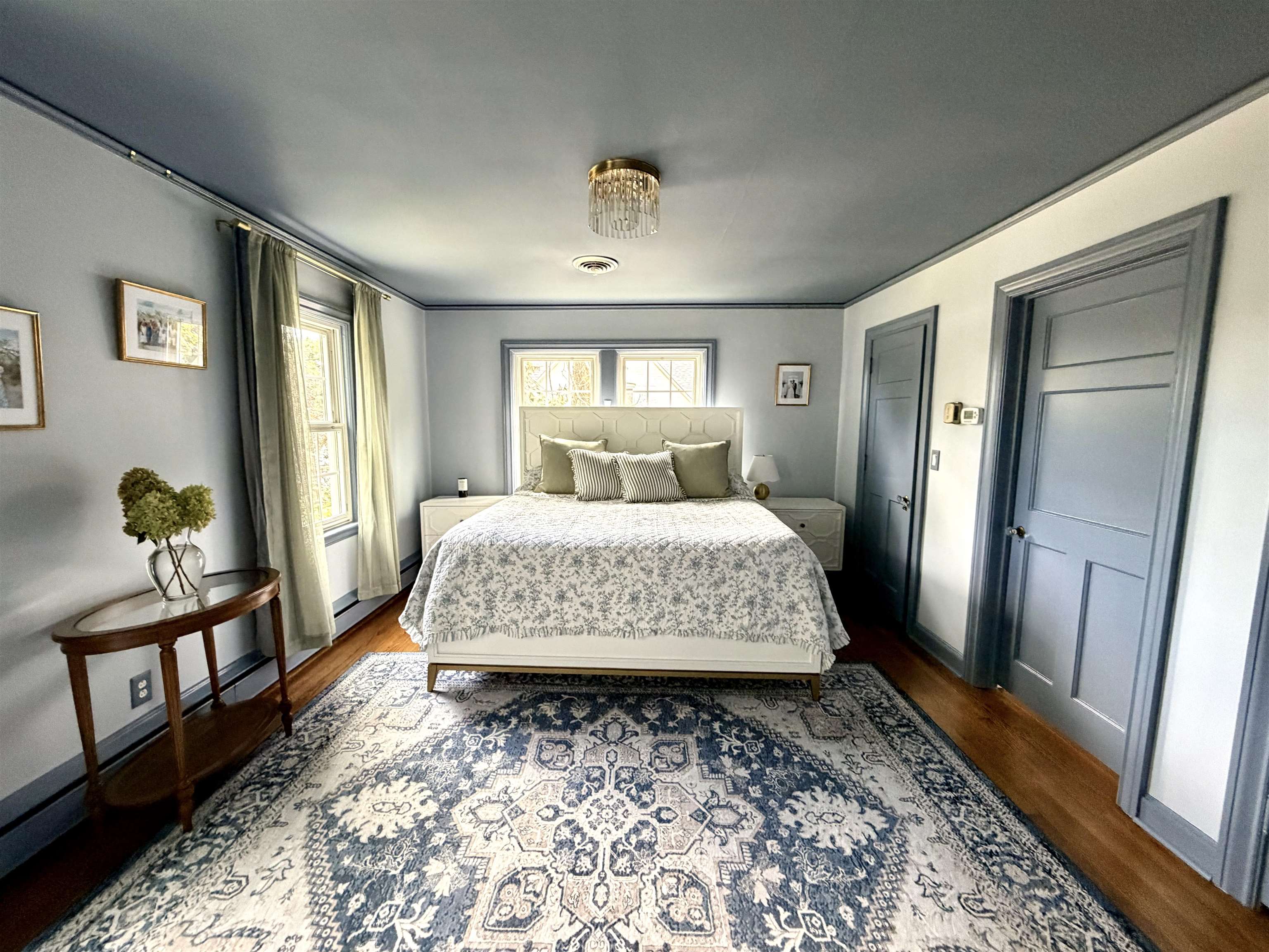 Bedroom with a centered bed, patterned rug, blue-gray trim, and tall curtained windows.