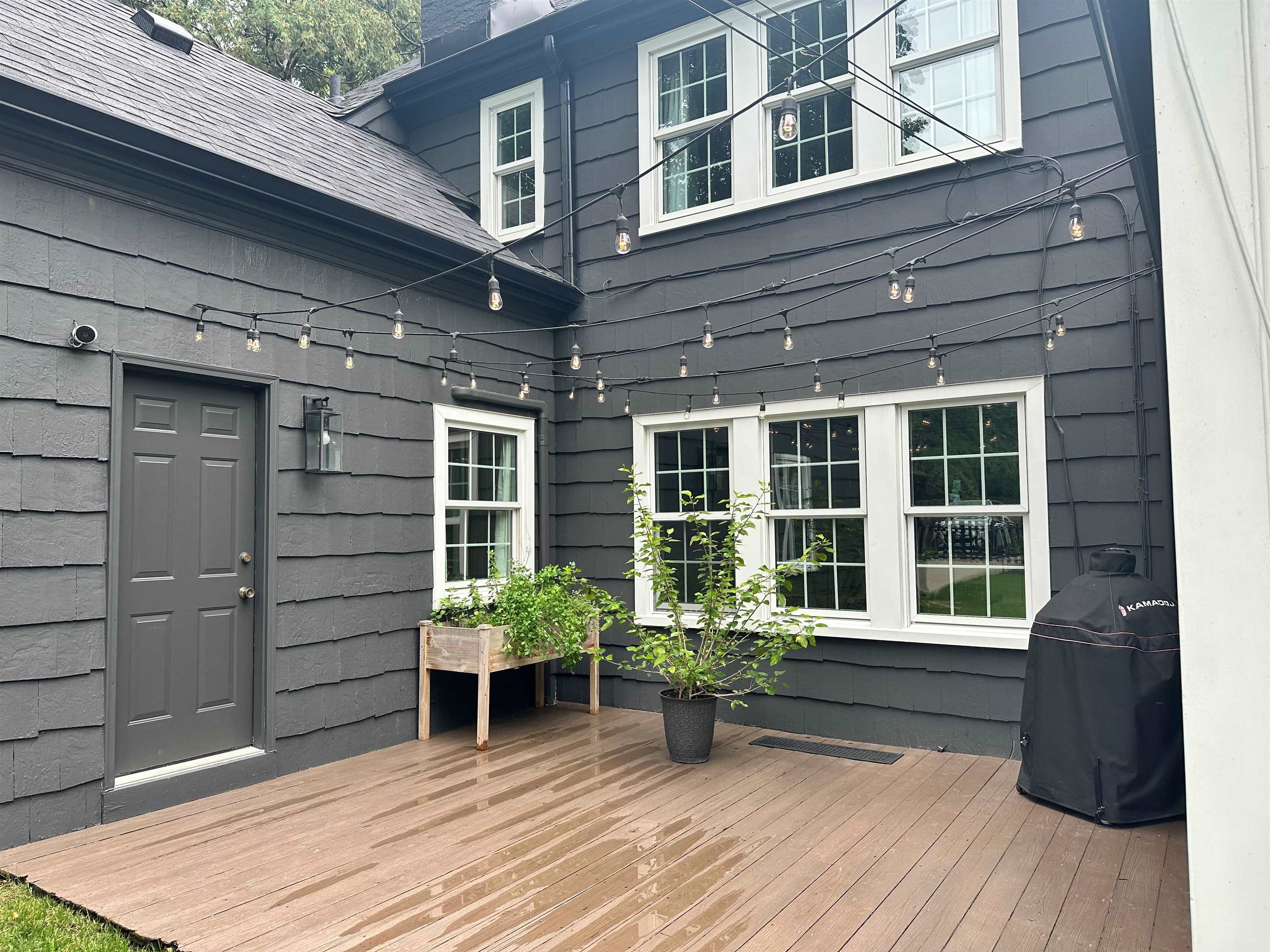 Wooden deck beside a dark gray house, with string lights, white-trimmed windows, and potted plants.