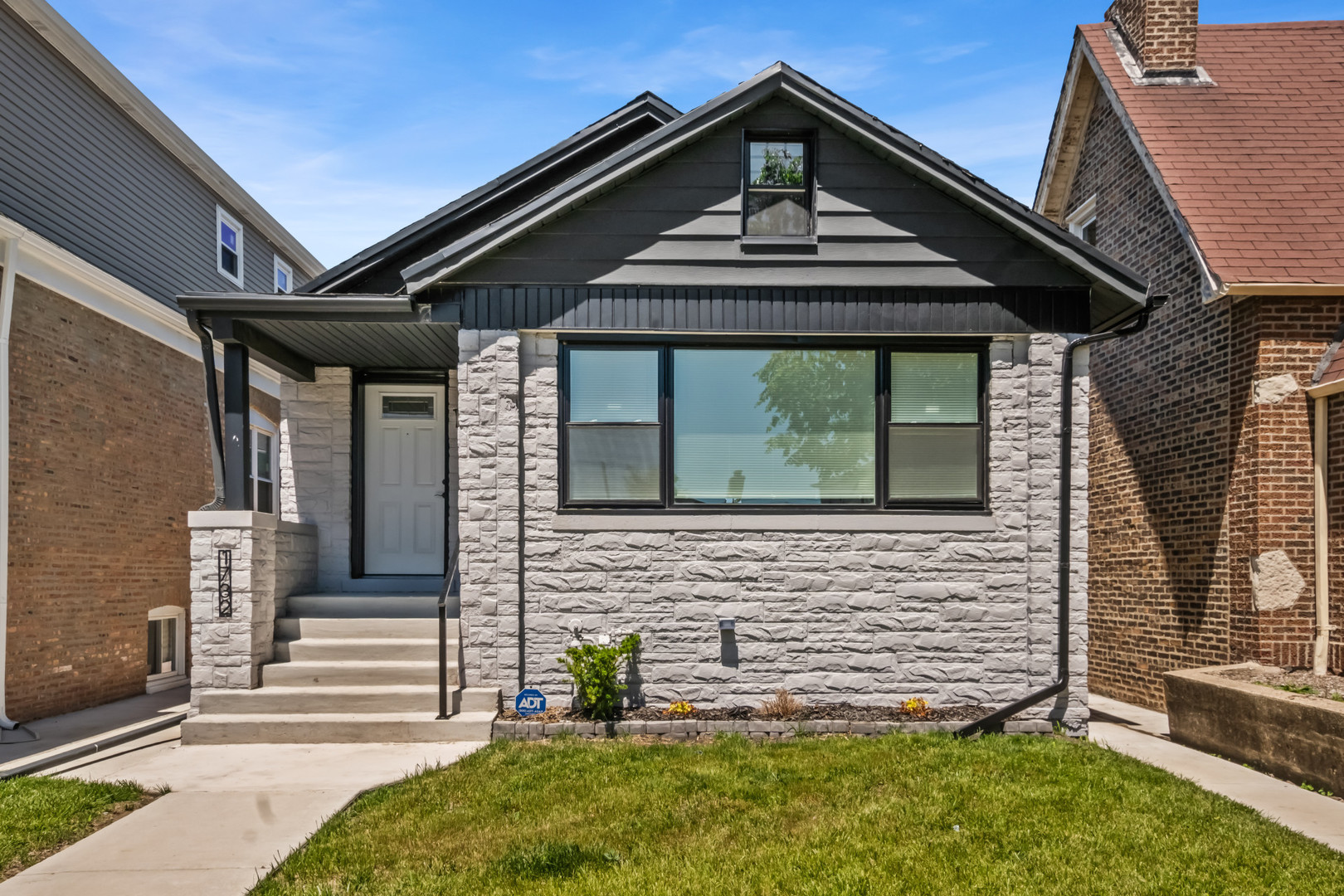 Gray stone-front house with a dark gabled roof, broad window, front steps, and a small lawn.