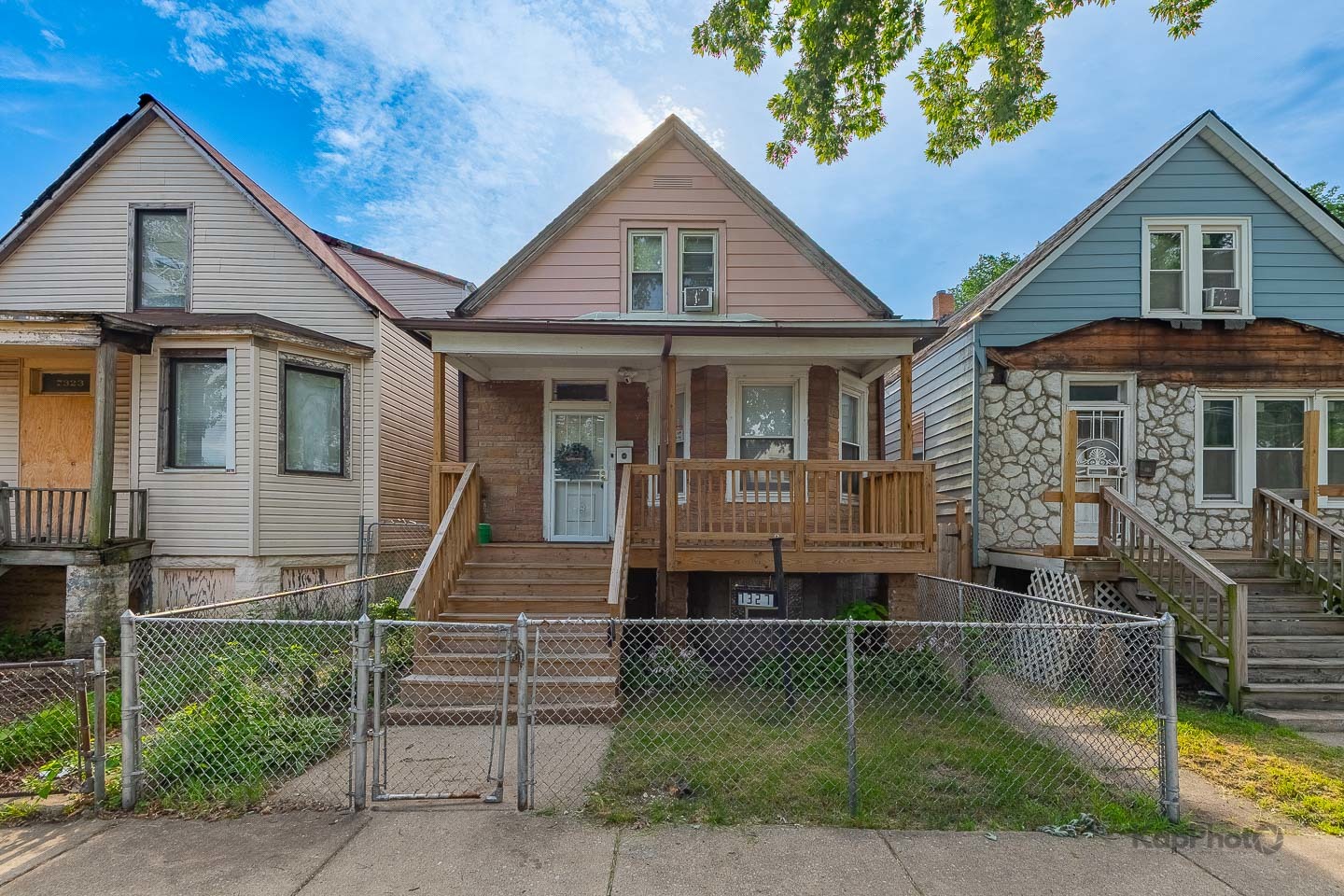 Gabled house with a raised wooden porch and chain-link fence, between two neighboring homes.