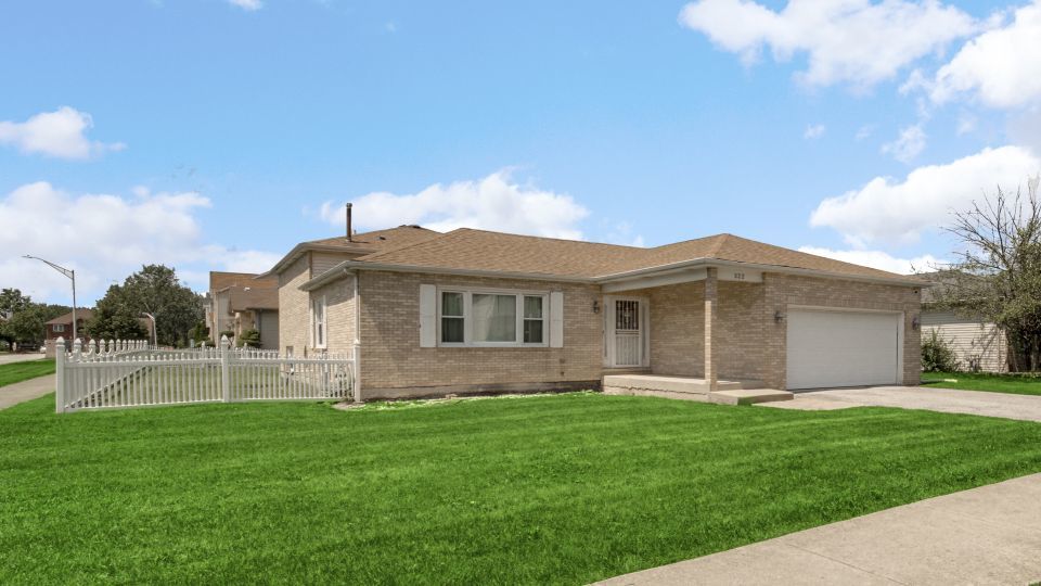 Beige brick ranch house with an attached garage, white picket fence, and broad green lawn.