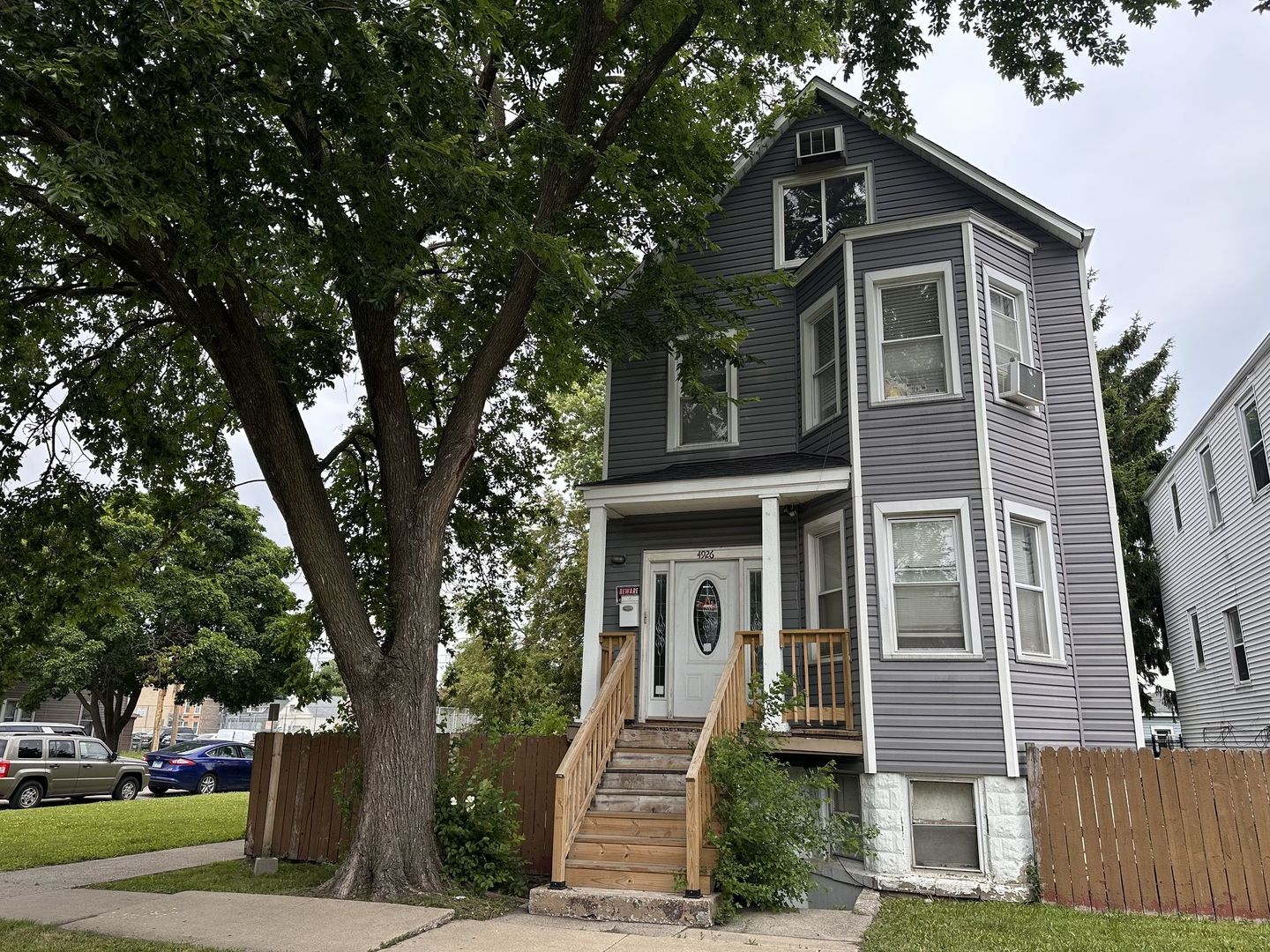Gray multi-story house with a raised wooden porch and large shade tree in the front yard.