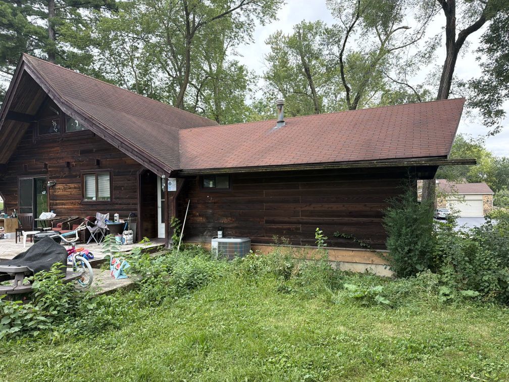 Dark wood-sided house with intersecting reddish roofs, set among trees and an overgrown lawn.