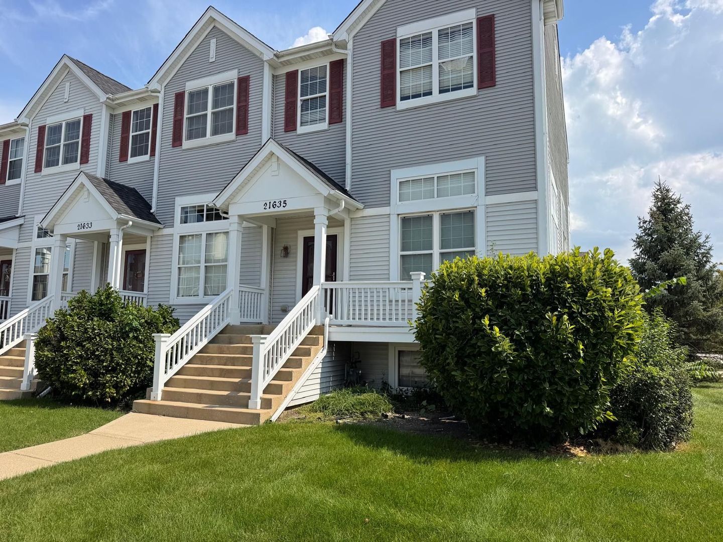 Gray-sided townhouse with red shutters, white porch railings, front steps, and trimmed shrubs.