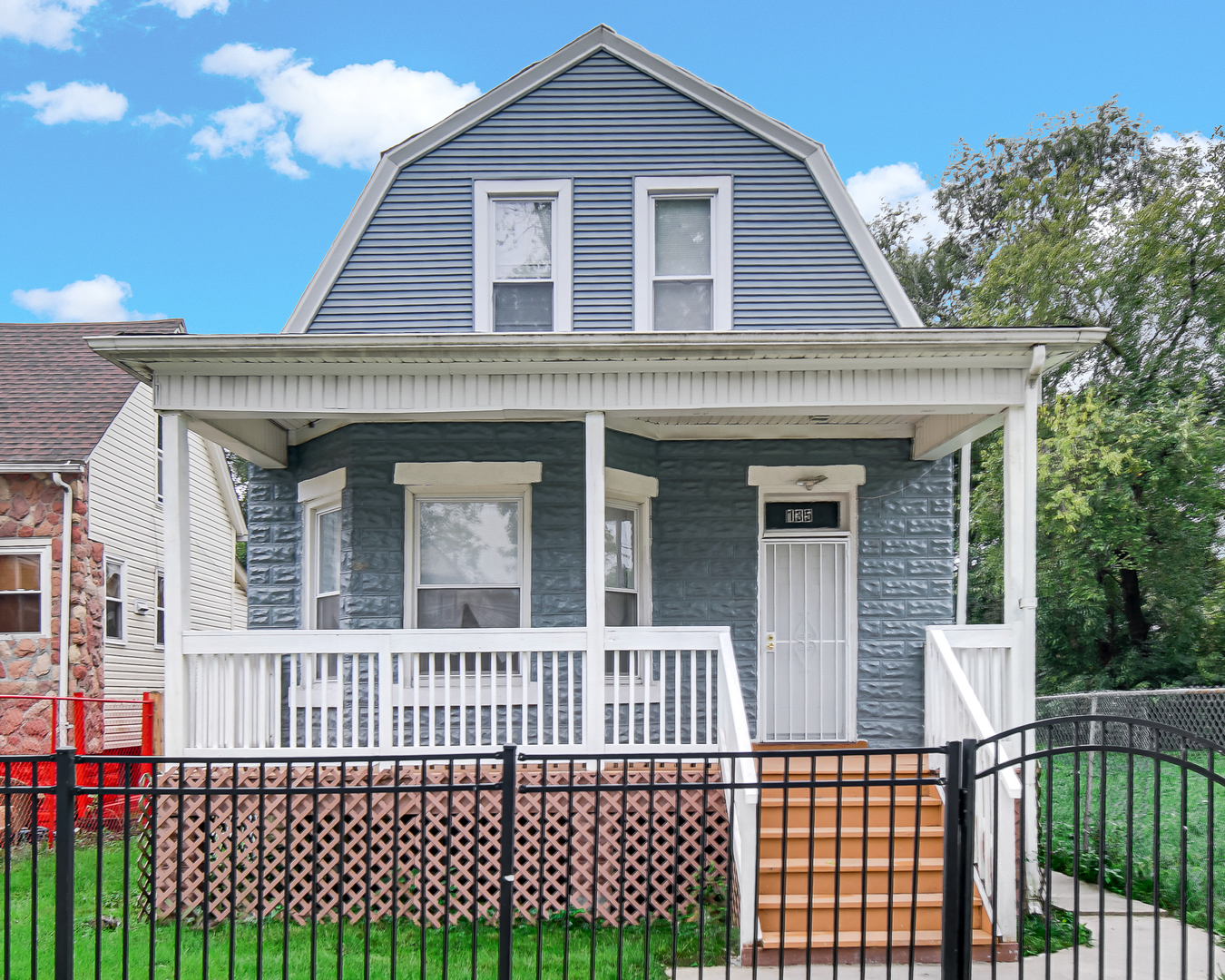 Blue-gray house with a steep gambrel roof, covered white porch, and black metal front fence.