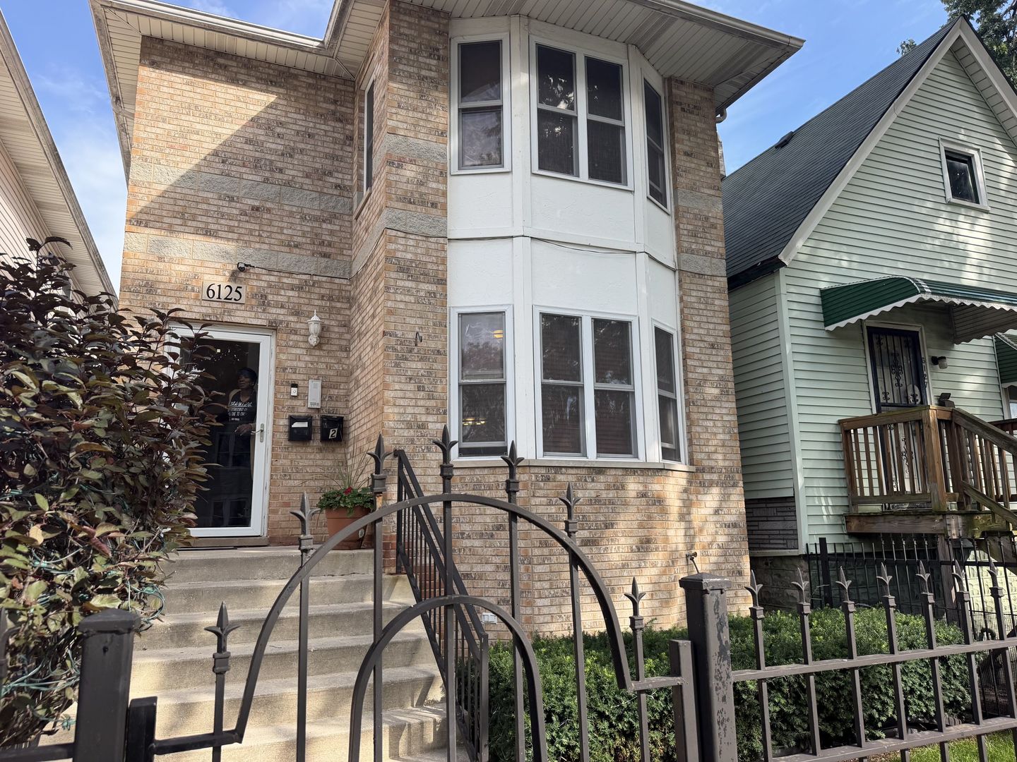 Tan brick house with a large white bay window, front steps, and black metal fence.