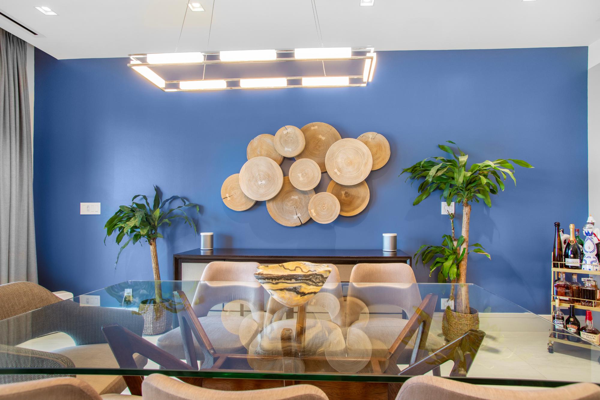 Dining room with a glass-top table, blue accent wall, circular wood art, and potted plants.
