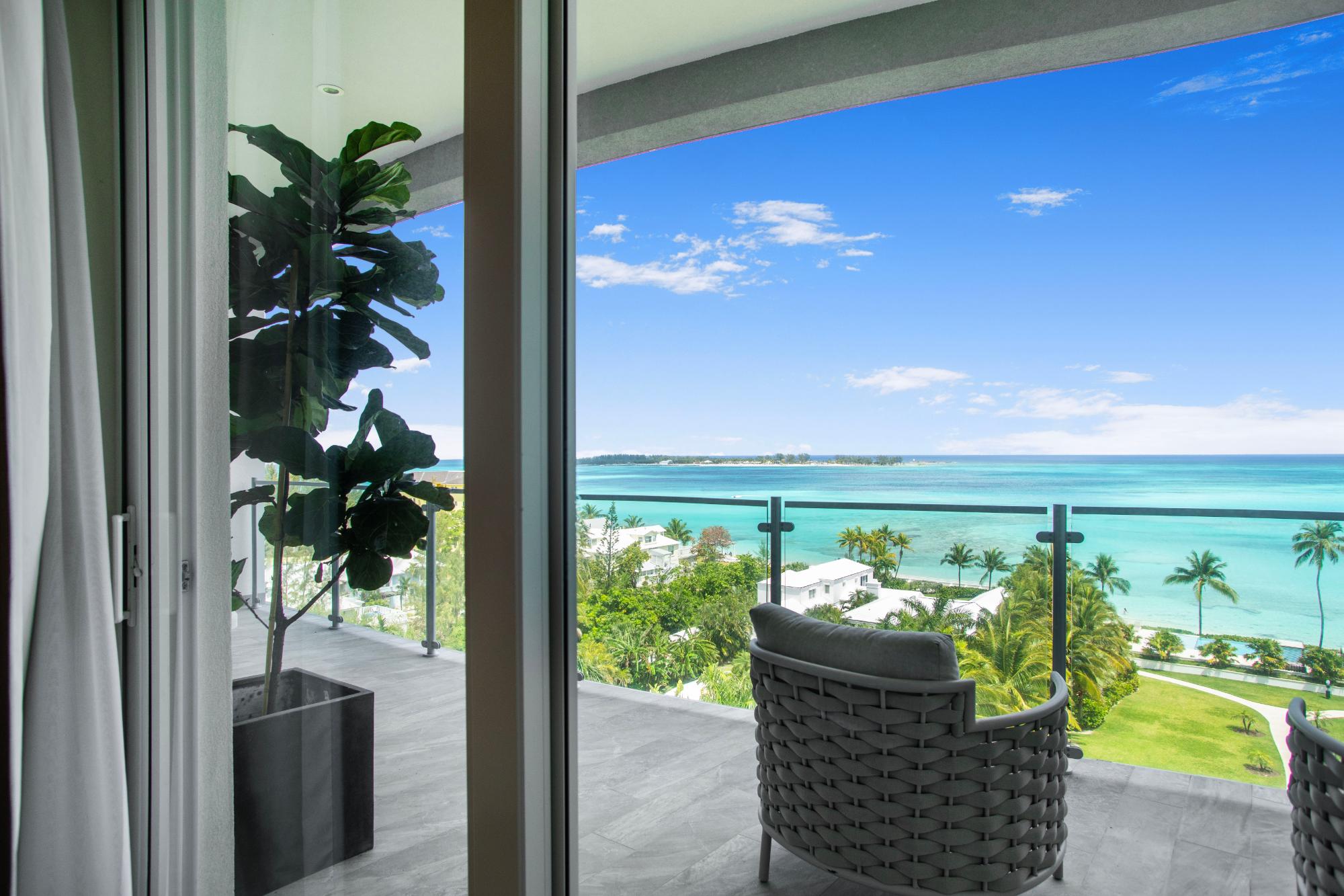 Furnished balcony overlooking turquoise ocean, with woven chairs and palm trees below.