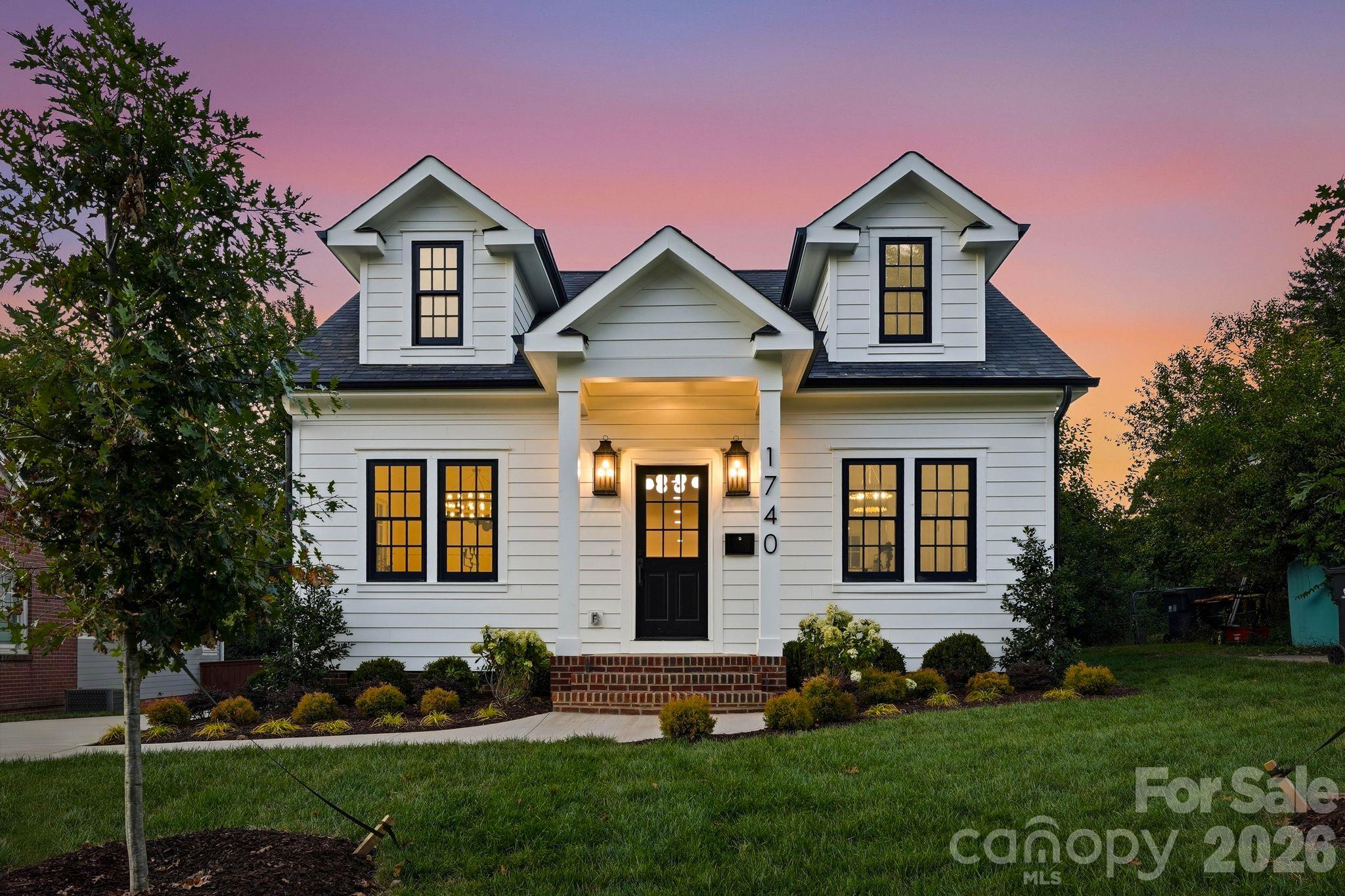 White two-story house with paired dormers and a warmly lit entrance at dusk.