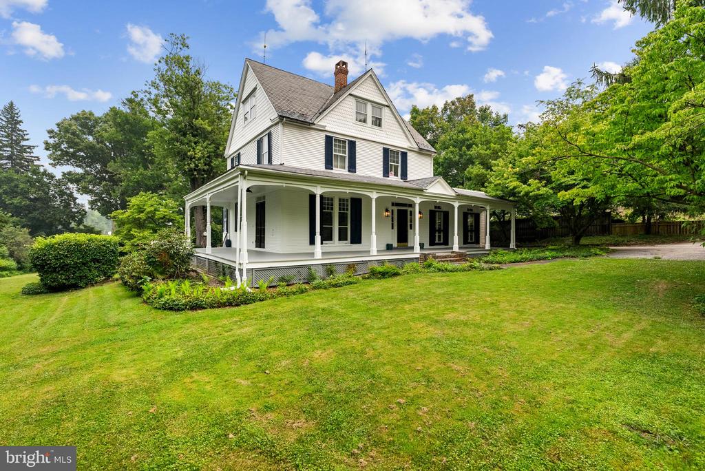 White two-story house with a wraparound porch, set behind a broad lawn and mature trees.