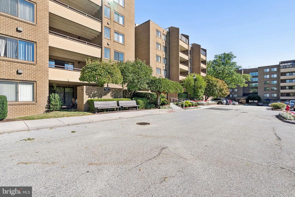 Brick condominium building with balconies, landscaped grounds, and benches beside the driveway.