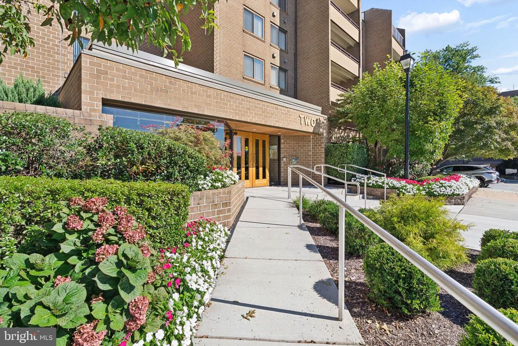 Brick residential building entrance marked TWO, approached by a ramp bordered with colorful flowers.