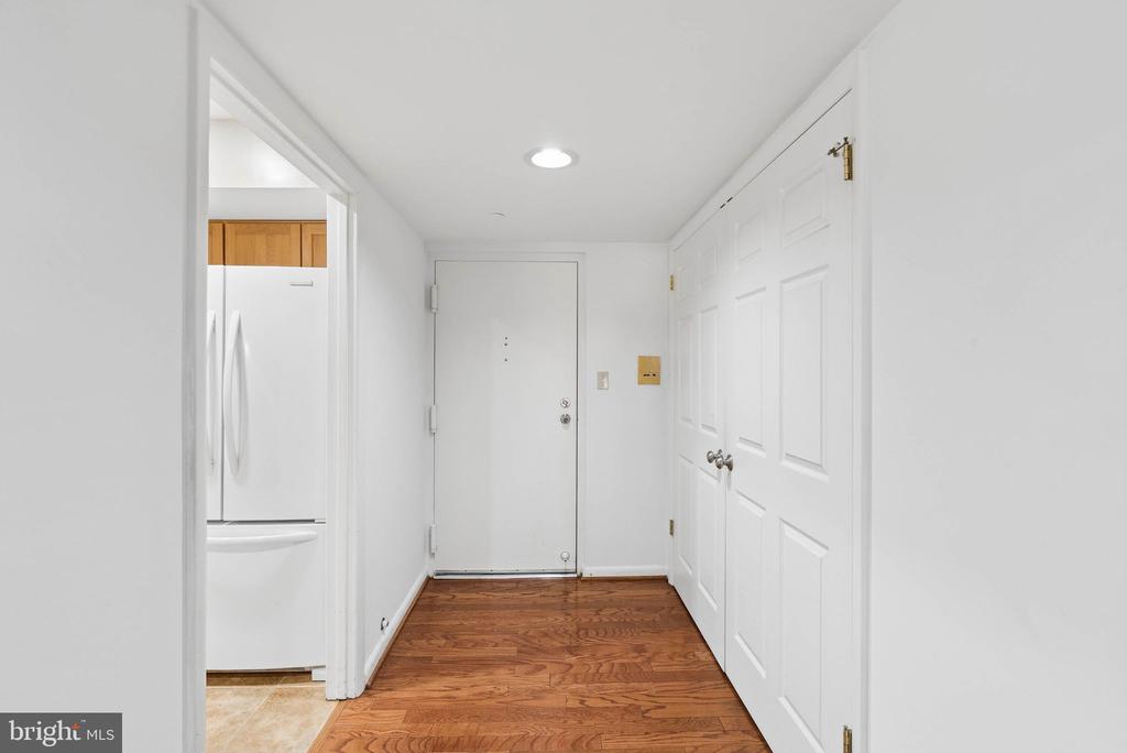Apartment entry hall with hardwood floors, paneled closet doors, and a refrigerator visible through the kitchen doorway.