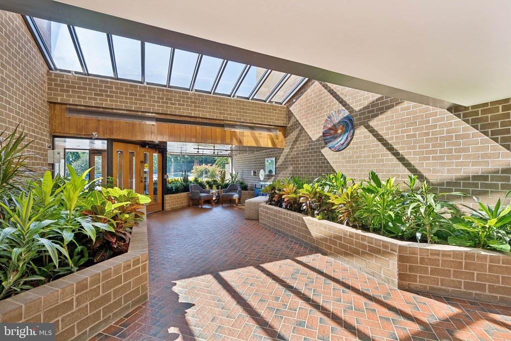 Sunlit brick lobby with a long skylight, tropical planters, and a small seating area.