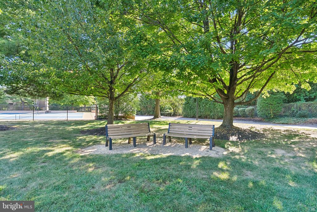 Community grounds with two benches beneath trees and a fenced swimming pool nearby.