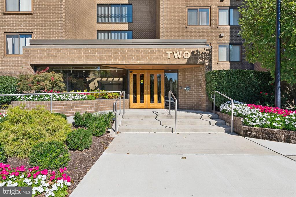 Brick apartment entrance with wood-framed glass doors, steps, metal railings, and colorful flower beds.