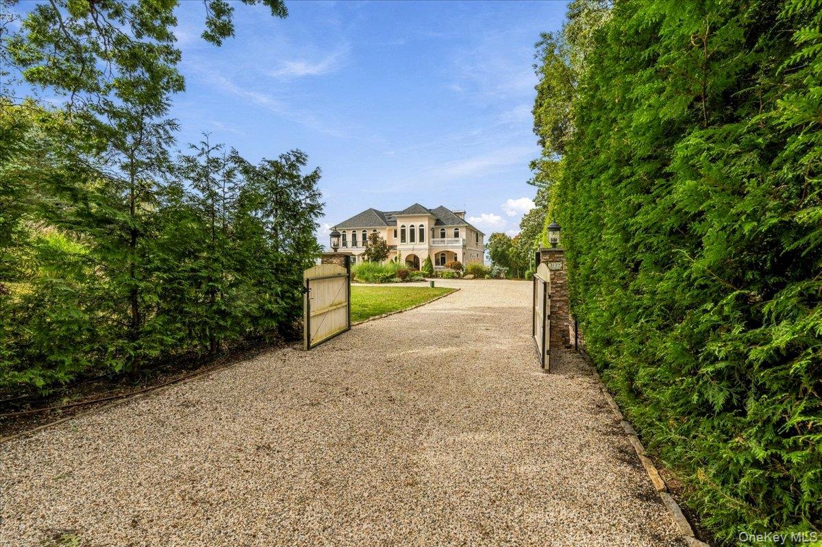 Large two-story home beyond an open gate at the end of a tree-lined gravel driveway.