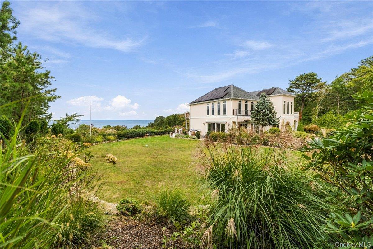 Two-story house on landscaped grounds overlooking the water, framed by trees and ornamental grasses.