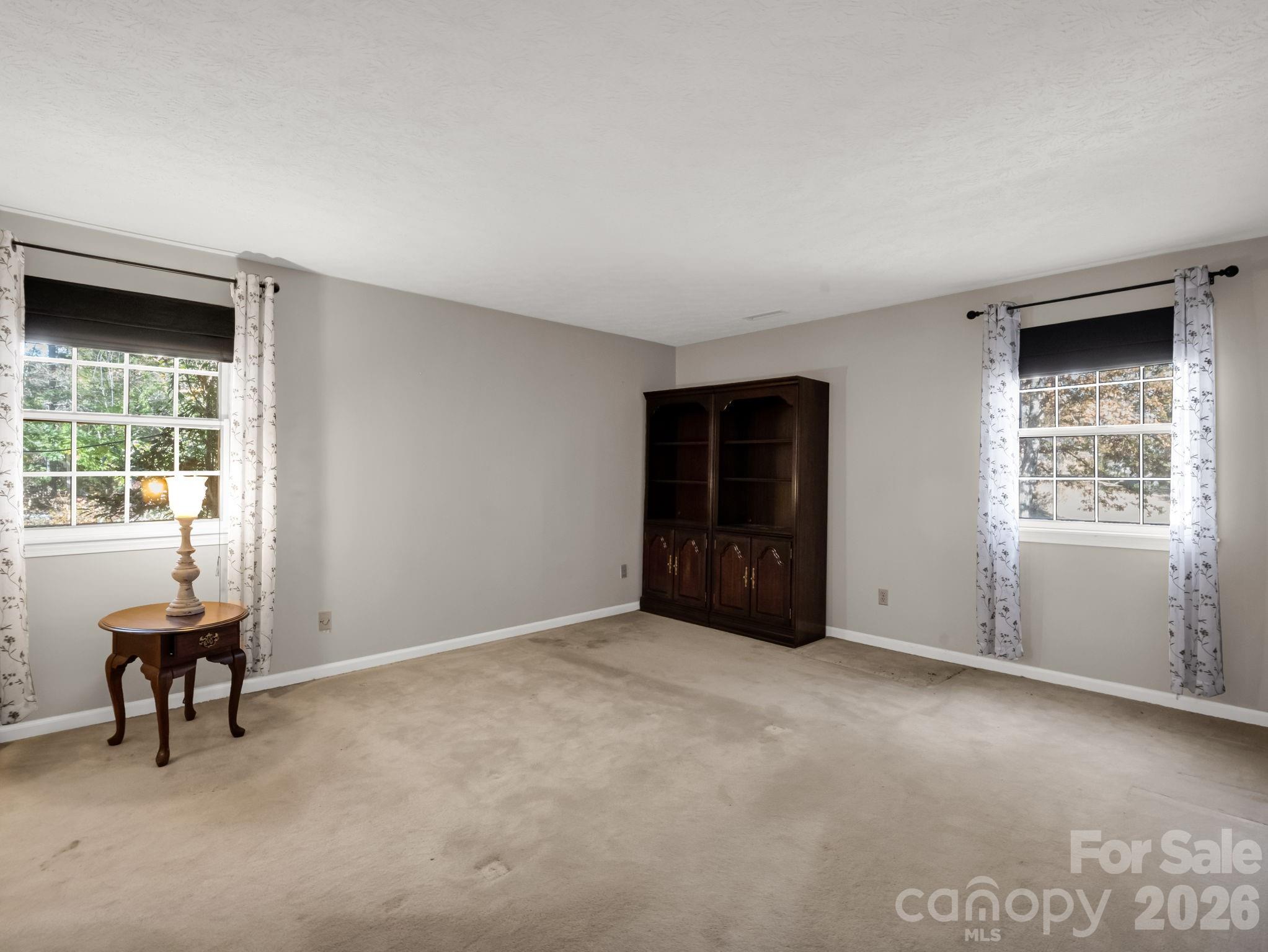 Carpeted room with two curtained windows, a dark bookcase, and a lamp on a small table.
