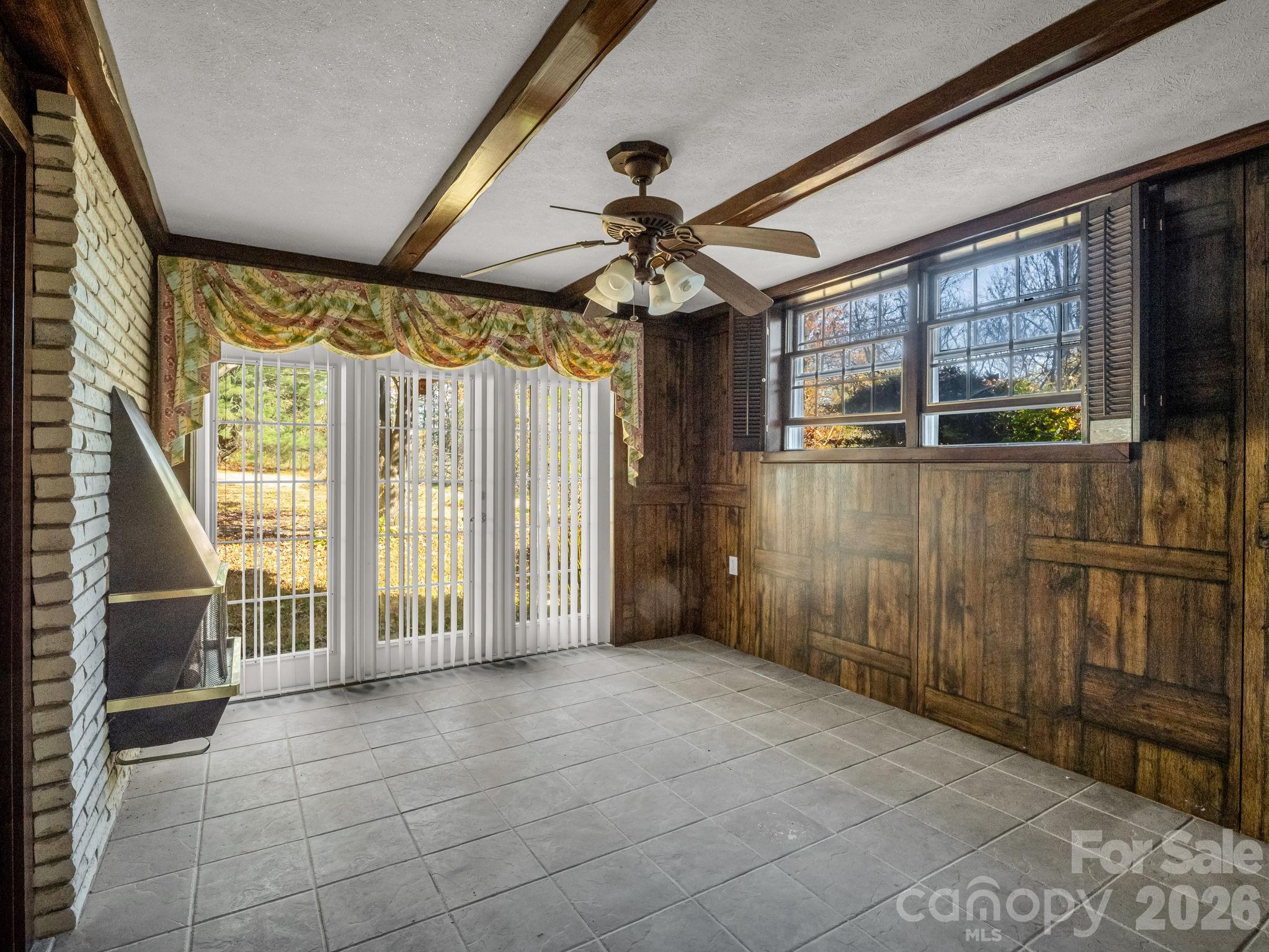 Empty tiled sunroom with dark wood paneling, exposed beams, and large windows with vertical blinds.